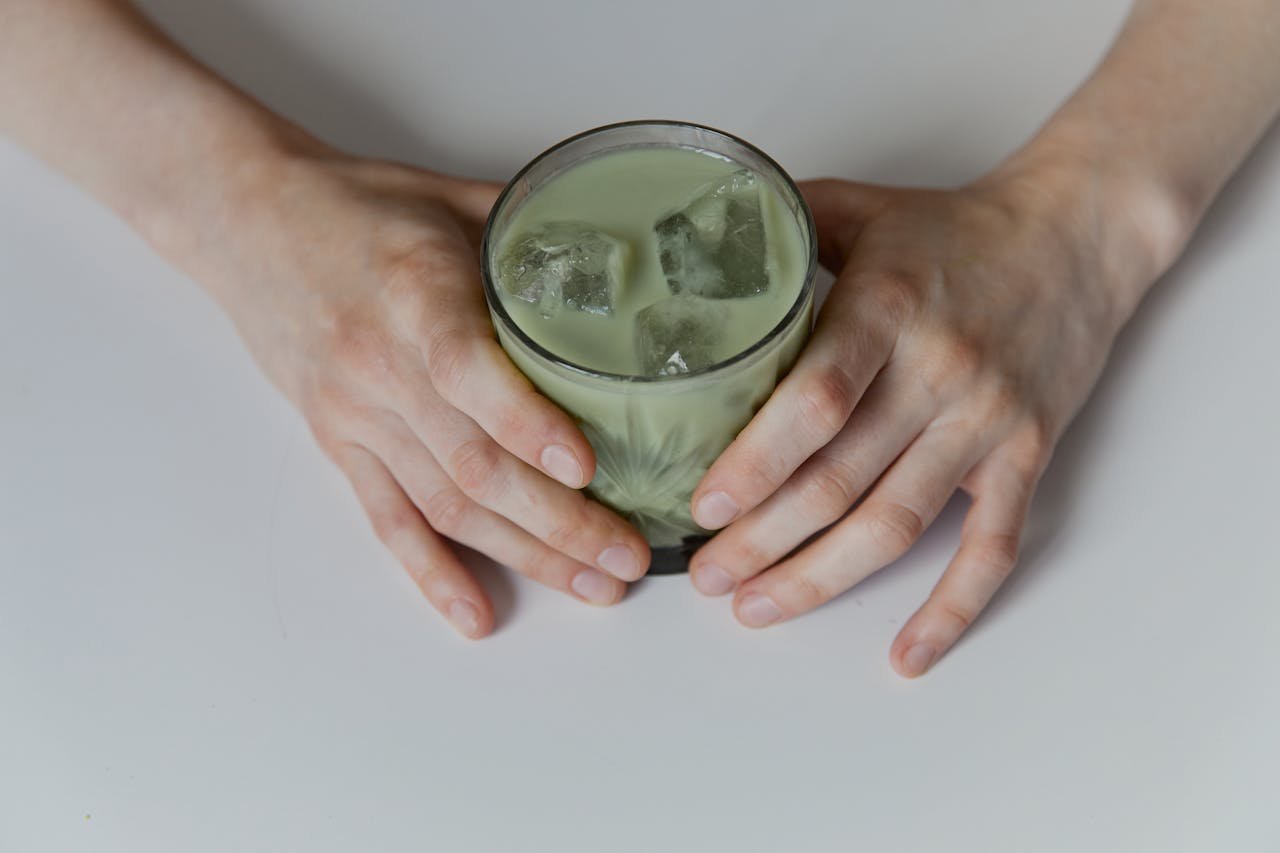 gallery-04 Close-up of a hand holding a glass of iced matcha latte on a white surface.