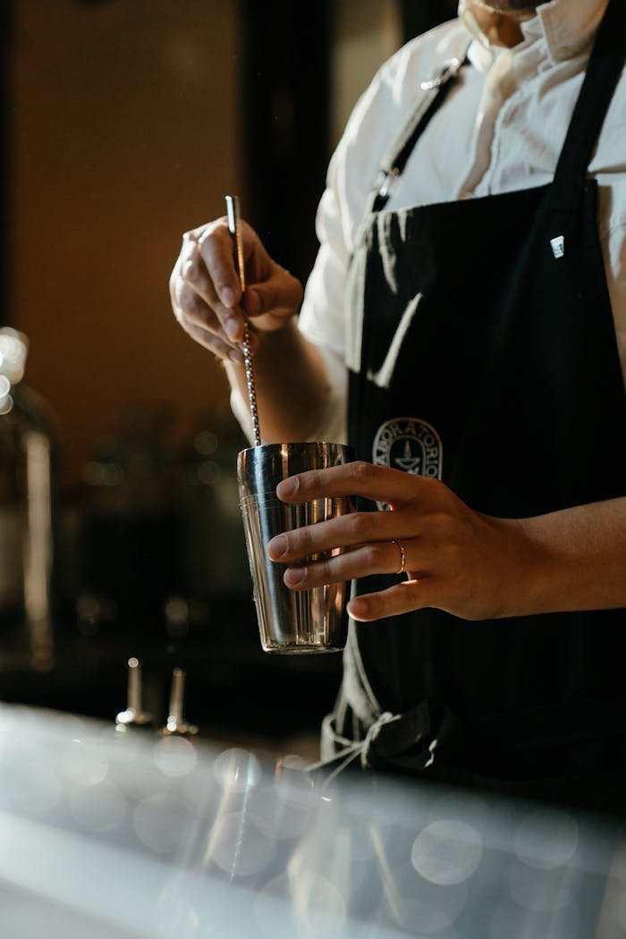 gallery-02 Close-up of a bartender stirring a cocktail with a bar spoon in a moody bar setting.